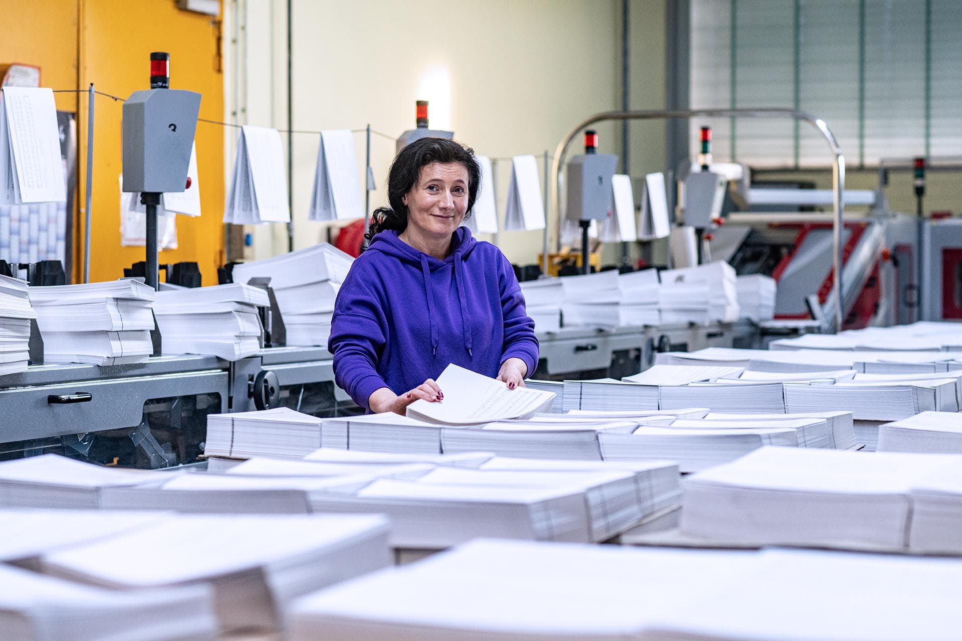 Eine sehr schöne Aufnahme mit starker Tiefenunschärfe einer freundlichen Mitarbeiterin mit blauem Hoodie, die liebevoll Buchblöcke für die Fadenheftung vorbereitet. Im Vorder- und Hintergrund sind unscharf unzählige Buchblöcke und die Produktionshalle mit Maschinen und Material der 