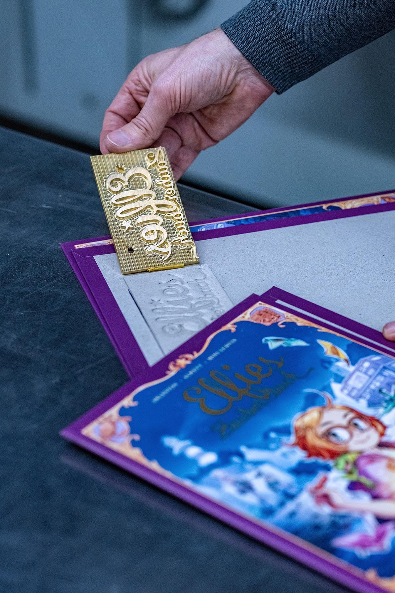 Eine sehr schöne Aufnahme mit starker Tiefenunschärfe einer Männerhand, die einen goldenen Prägestempel mit geschwungener Schrift hält. Auf einem Tisch liegt ein geprägter Einband mit Prägung von hinten gesehen, davor ist unscharf der geprägte bunte Einband von vorne zu sehen. Im Hintergrund ist unscharf Produktionshalle der 